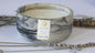 A candle with a gray marbled container, the label reads 'Promised Land' with a description of the scent. The candle is accompanied by some eucalyptus leaves and there is a lit candle in the background.