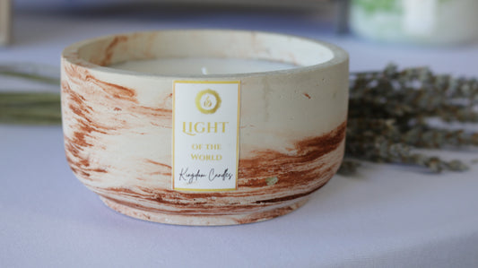 A candle with a beige and brown marbled-like pattern on the top, labeled 'Light of the World', with a bronze color wax pool visible.
